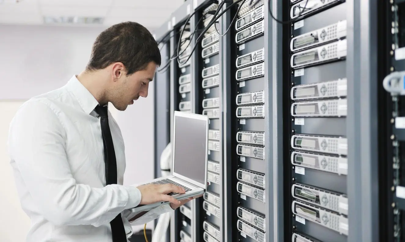 A man in white shirt using laptop near server racks.